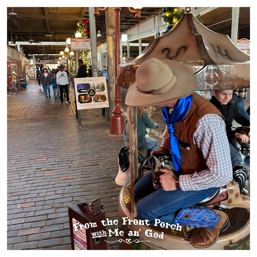 Acowboy sits on a carousel at what appears to be a mall. A text overlay on the image says "From the Front Porch with Me an' God".